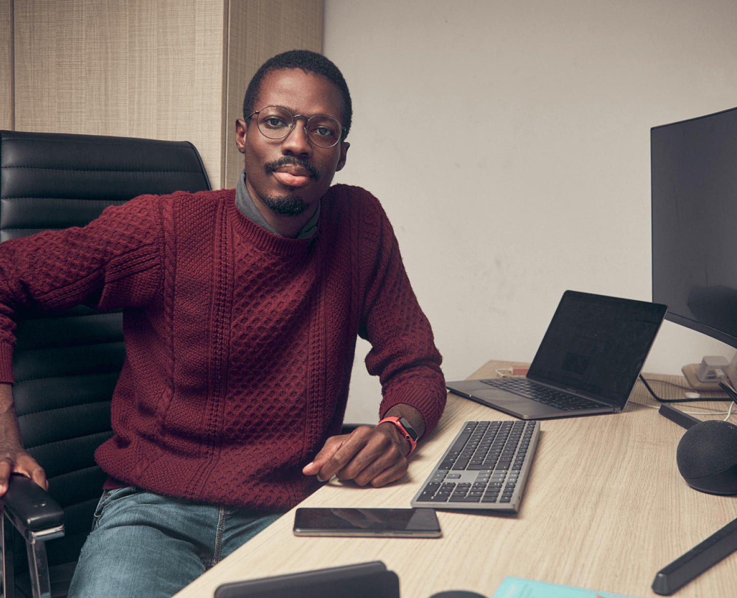 A man wearing a maroon knit sweater, sitting on an office chair and resting an arm on his work desk while staring into the camera.