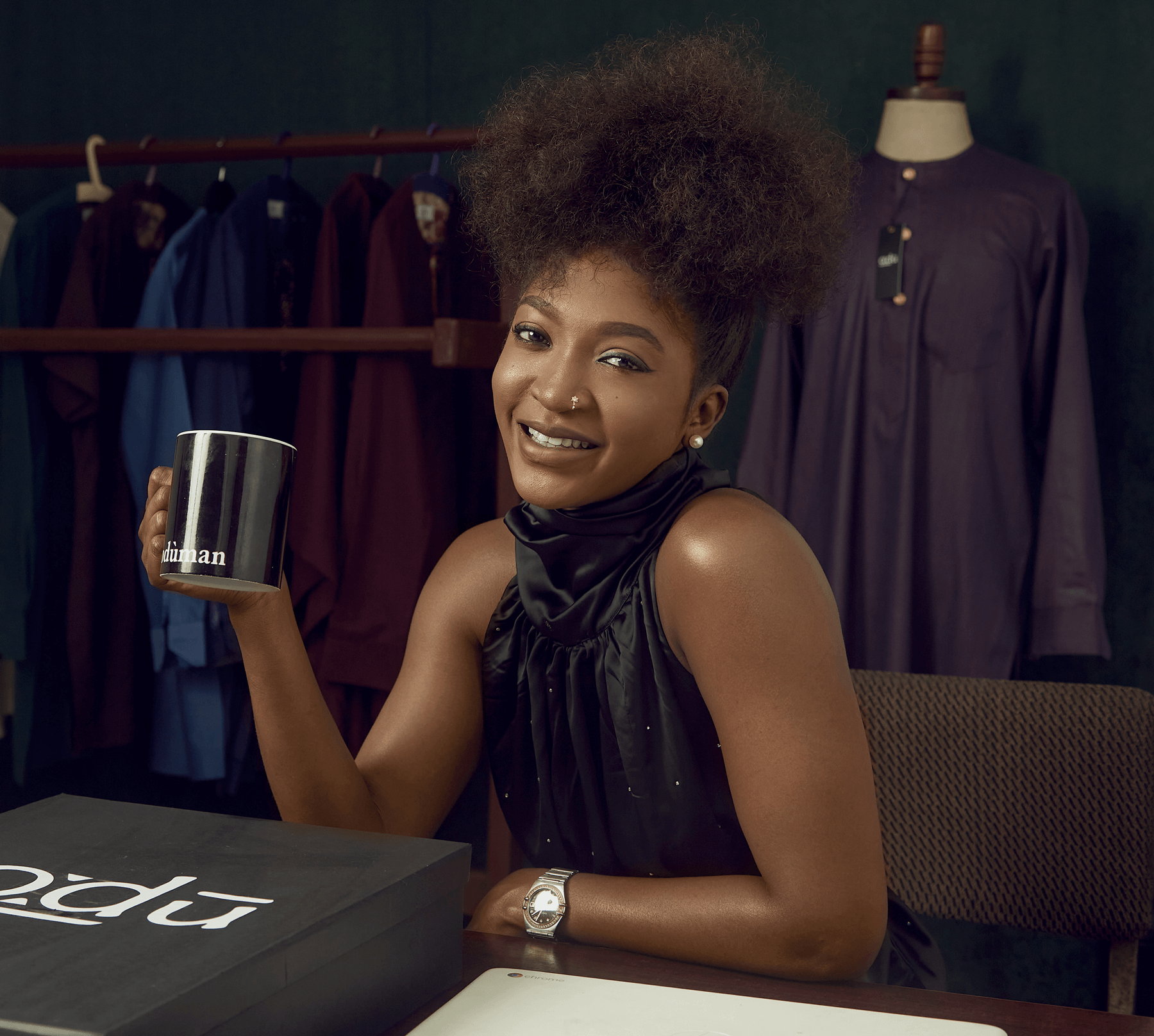 A woman holding a cup, sitting on a chair while resting an arm on her desk. There is a coat rack behind her as she smiles into the camera.